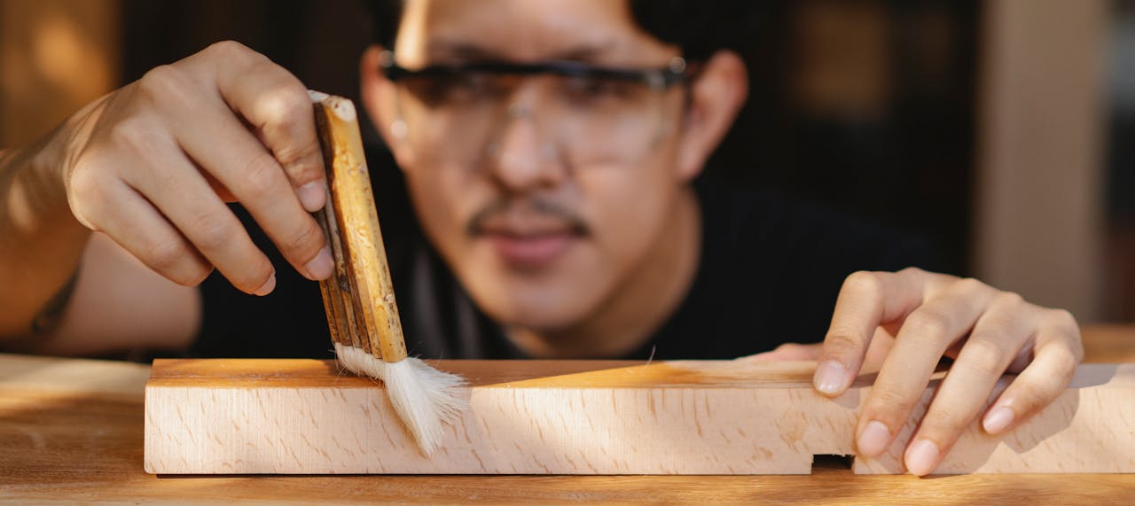 Young Asian craftsman in protective eyeglasses applying glue on wooden stick while working in professional workshop